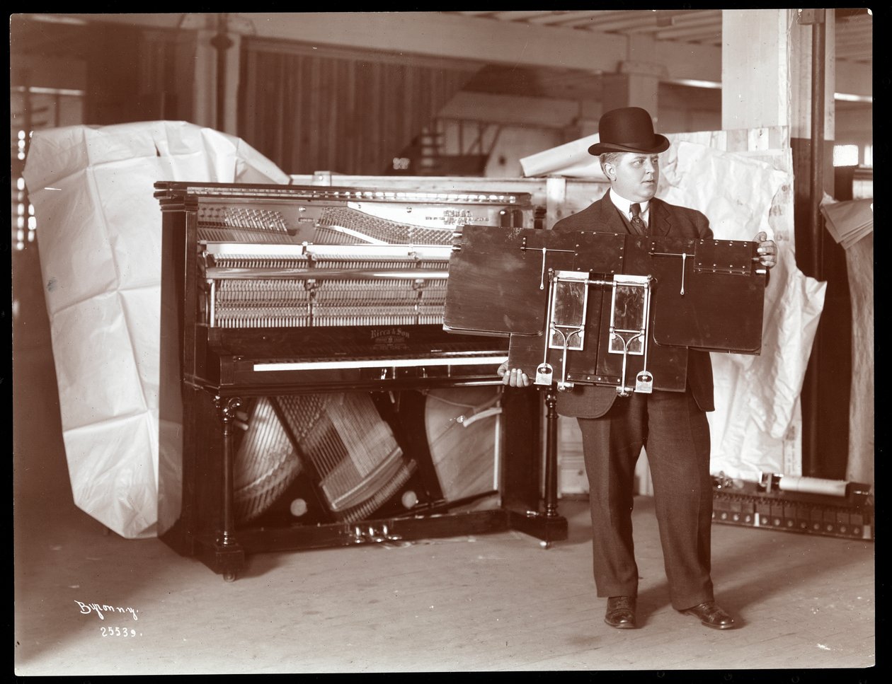Fortepian firmy Ricca and Son Piano Co., obok którego mężczyzna podtrzymuje część utworów fortepianowych, 1907 (srebrna odbitka żelatynowa) autorstwa Byron Company