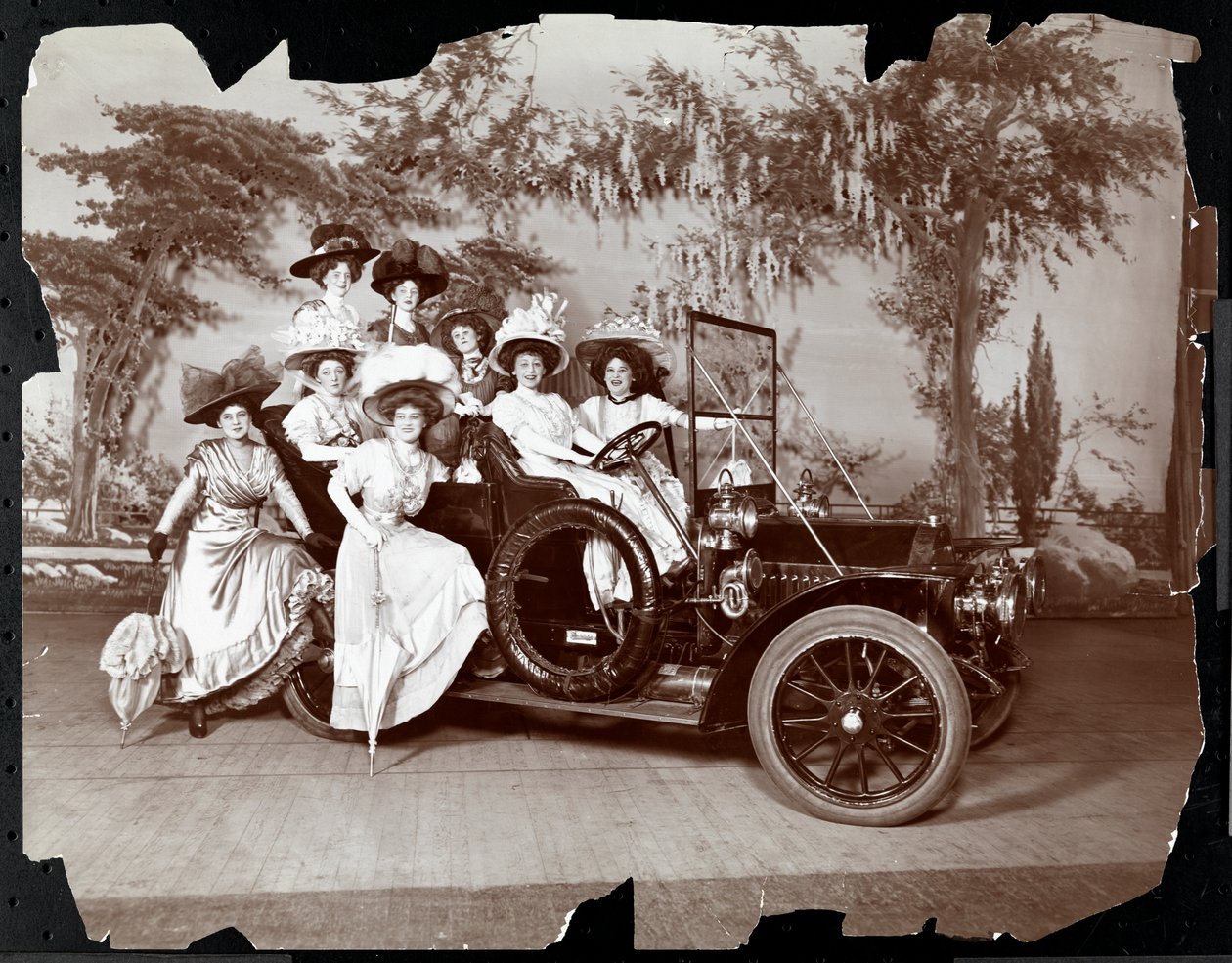 Osiem kobiet w kostiumach pozowało w samochodzie i wokół niego na scenie z krajobrazem, prawdopodobnie w przedstawieniu „The Vanderbilt Cup”, 1906 (srebrny odbitka żelatynowa) autorstwa Byron Company