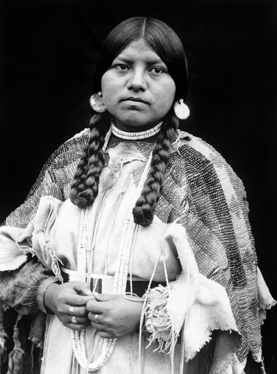 Kobieta Cayuse, portret w połowie długości, stojąca, zwrócona do przodu, warkocze, kolczyki w kształcie dysków muszli, naszyjnik z muszelek i koraliki muszli na szyi, suknia ze skóry bydlęcej z koralikami i paskiem z koralików c. 1910, fot. Edward S. Curti autorstwa Edward Sheriff Curtis