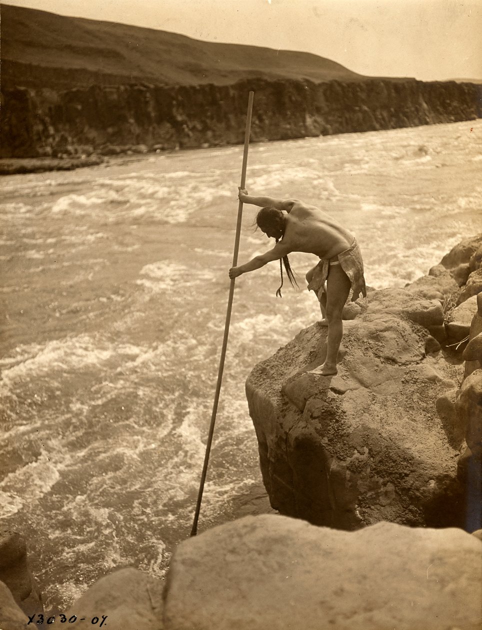 Indianin Tlakluit, stojący na skale, łowiący ryby siecią, ok. 1910 r. (zdjęcie czarno-białe) autorstwa Edward Sheriff Curtis
