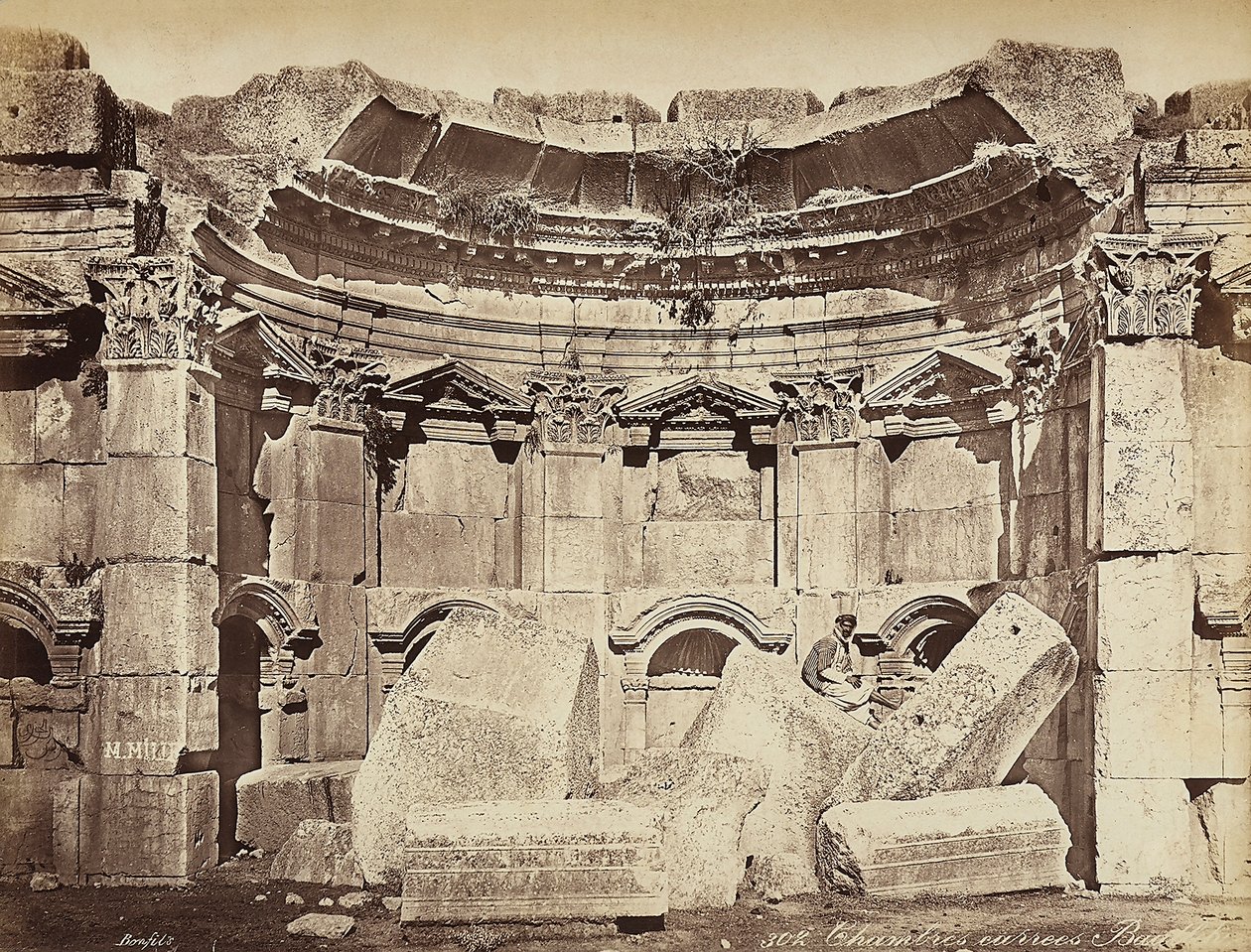 Fragment tzw. „pokojów” w strefie archeologicznej Heliopolis lub Baalbek, starożytnego syryjskiego miasta, obecnie Libanu. Mężczyzna pozuje, siedząc na ruinach autorstwa Félix Bonfils