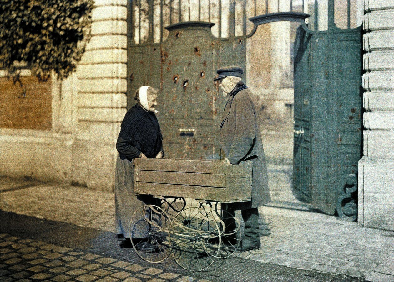 Dwoje starszych mieszkańców Reims, starsza kobieta i pan Reiser, stoją przed bramą z wieloma dziurami po kulach, Reims, Marna, Francja, 1917 (autochrom) autorstwa Fernand Cuville