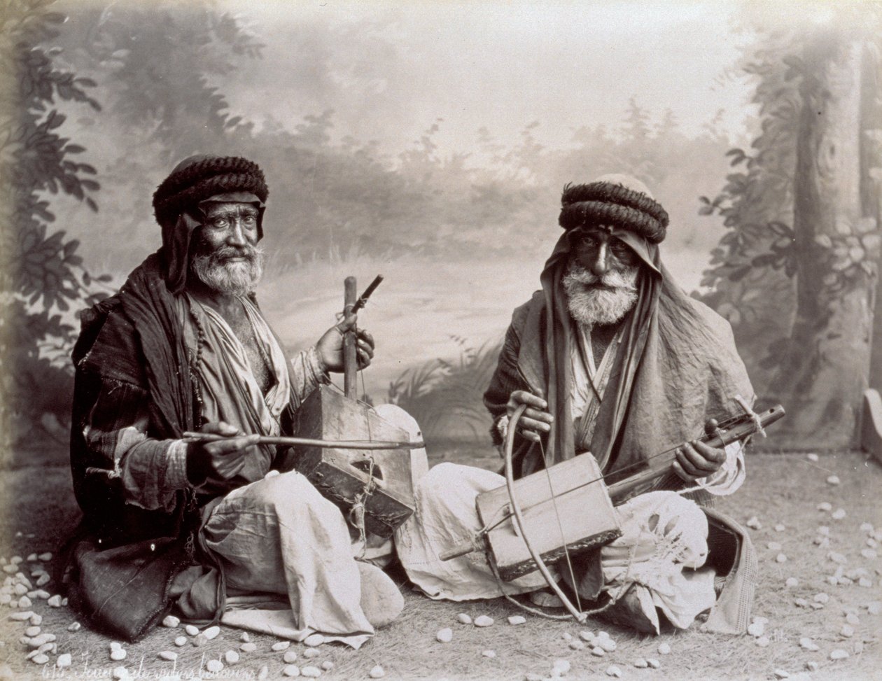 Portret dwóch arabskich muzyków w skromnych etnicznych strojach. Starsi panowie pozują w studio, siedząc na ziemi. Za nimi widać zalesiony krajobraz autorstwa Félix Bonfils
