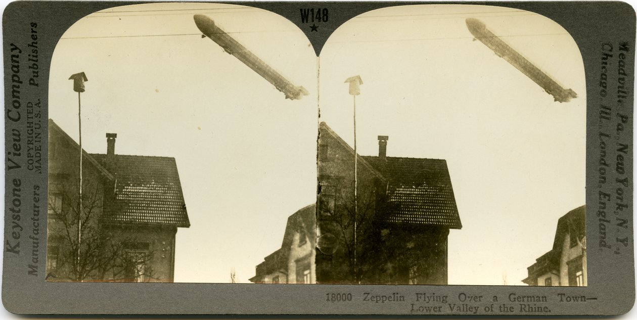 Pozioma karta stereofoniczna w sepii przedstawiająca Zeppelina przelatującego nad dachami miasta. Keystone Stereograph numer 18000. Tytuł brzmi autorstwa Keystone View Company