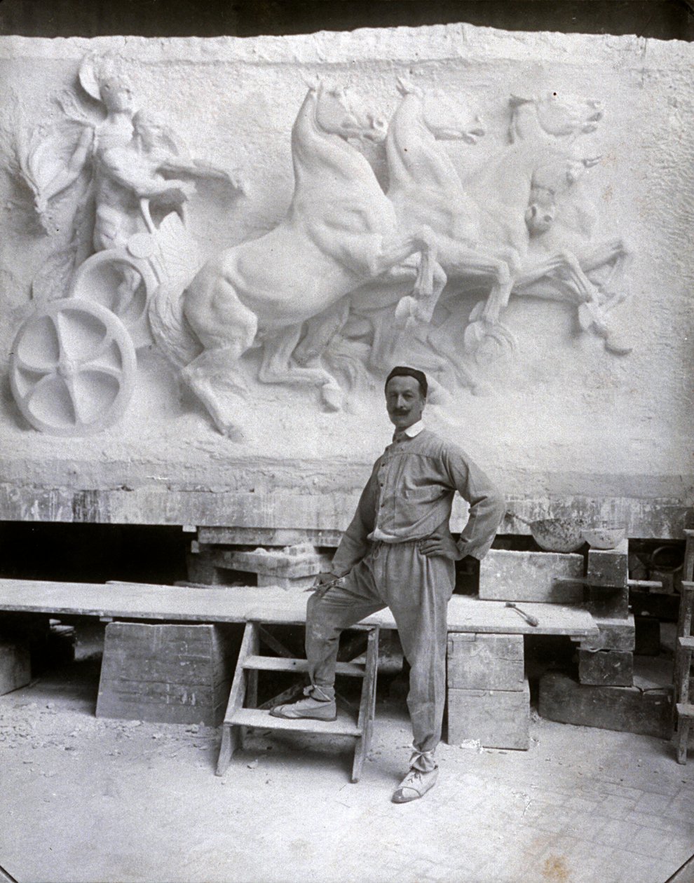 Pełnowymiarowy portret słynnego rzeźbiarza Pietro Canonica. Pozuje w swoim studio przed marmurową płaskorzeźbą, nad którą pracuje (zdjęcie czarno-białe) autorstwa Mario Nunes Vais