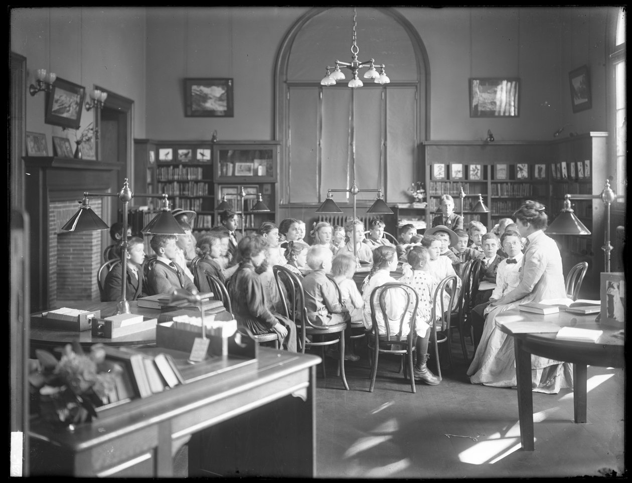 Dzieci słuchają kobiety w czytelni niezidentyfikowanej filii Biblioteki Publicznej Queens Borough, ok. 1910 autorstwa William Davis Hassler