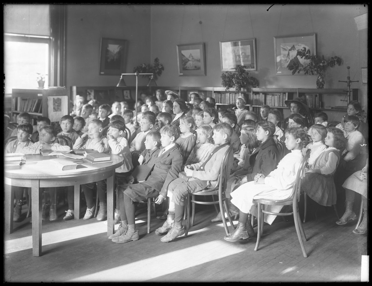 Dzieci siedzące, słuchające w czytelni niezidentyfikowanej filii biblioteki publicznej w Queens Borough, ok. 1910 r. autorstwa William Davis Hassler