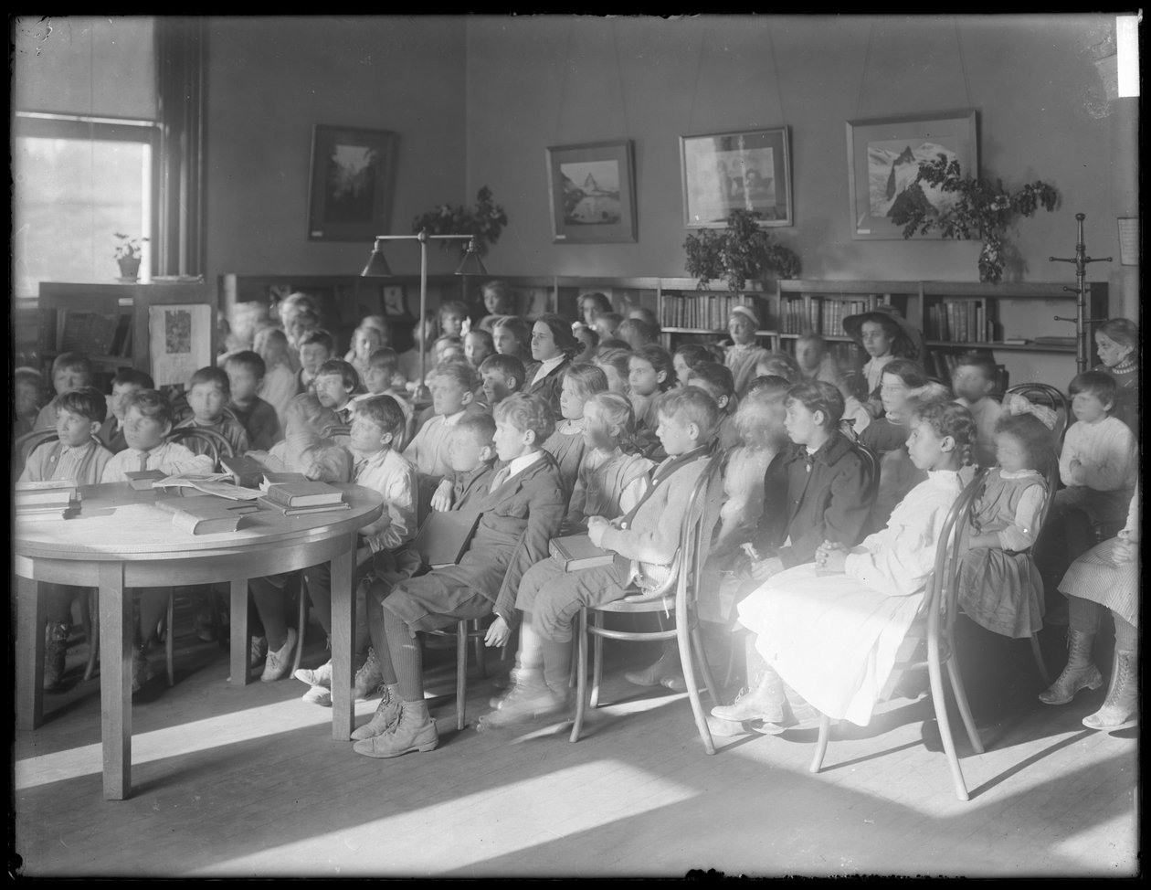Dzieci siedzące, słuchające w czytelni niezidentyfikowanej filii biblioteki publicznej w Queens Borough, ok. 1910 r. autorstwa William Davis Hassler