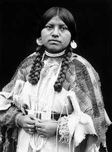 Kobieta Cayuse, portret w połowie długości, stojąca, zwrócona do przodu, warkocze, kolczyki w kształcie dysków muszli, naszyjnik z muszelek i koraliki muszli na szyi, suknia ze skóry bydlęcej z koralikami i paskiem z koralików c. 1910, fot. Edward S. Curti autorstwa Edward Sheriff Curtis