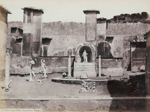 Częściowy widok na podwyższony ogród Domu Marka Lukrecjusza w Pompejach, ozdobiony marmurowymi posągami i hermami. W centrum fontanna z wnęką. W tle fragmenty malowideł ściennych autorstwa Giorgio Sommer
