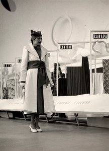 Model w płaszczu stworzonym przez dom mody "Lamma". Artykuł jest zebrany w talii szeroką szarfą i wykończony miękkim kapeluszem. Zdjęcie zostało zrobione w sali przeznaczonej na ekspozycję tkanin. (zdjęcie czarno-białe) autorstwa Luis Bogino
