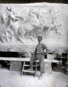 Pełnowymiarowy portret słynnego rzeźbiarza Pietro Canonica. Pozuje w swoim studio przed marmurową płaskorzeźbą, nad którą pracuje (zdjęcie czarno-białe)