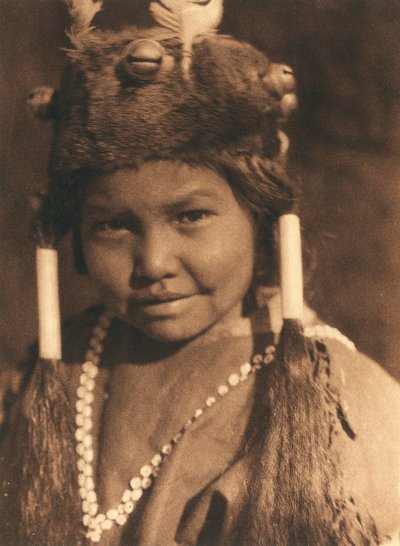 Zbliżenie na amerykańskiego Indianina z plemienia lub wioski Klamath. Ma na sobie futrzaną czapkę z aplikacjami z żołędzi i piór, a włosy nosi spięte w dwa kucyki autorstwa Edward Sheriff Curtis