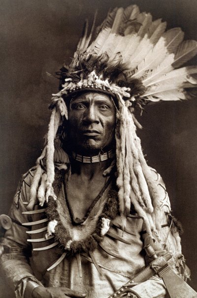 Ogon łasicy (Apohsuyis). Portret wodza Indian z plemienia Piegan ma na sobie słynne pióro orła i nakrycie głowy łasicy, zamszową tunikę, dwa naszyjniki, jeden z kości, a drugi z pazurami grizzly, oraz fajkę tomahawk autorstwa Edward Sheriff Curtis
