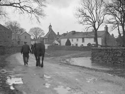 Mężczyzna prowadzący konia drogą z dala od kamery w kierunku budynków wiejskich i kościoła; architektura; religijny; kościelny; autorstwa Joseph Hardman