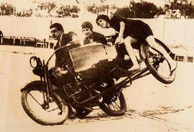 Mężczyzna popisuje się jazdą na motocyklu z wózkiem bocznym przewożącym trzy młode kobiety. Wózek boczny podnosi się z ziemi, gdy wykonują zakręt. autorstwa Unknown photographer