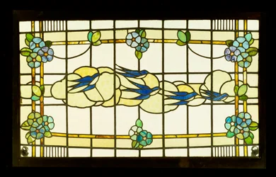 Panel Arts and Crafts przedstawiający grupę jaskółek przed chmurami w obramowaniu stylizowanych główek kwiatów (ołów i witraż)