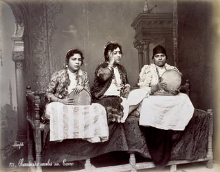Portret trzech młodych arabskich śpiewaków w etnicznych strojach z długimi naszyjnikami. Pozują w studio, siedząc. Dwóch z nich trzyma tamburyn