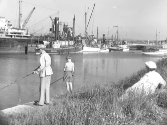 Mężczyzna i chłopiec łowią ryby w Glasson Dock, a siedząca za nimi dama przygląda się, kilka łodzi jest zacumowanych