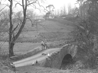 Dwie dziewczynki siedzące na kamiennym moście nad rzeką Cocker u podnóża Scale Hill, obserwujące gęsi na drodze w Loweswater