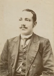 Zdjęcie mężczyzny w ciemnym garniturze z kieszonkowym zegarkiem w kamizelce, 1886-1900.