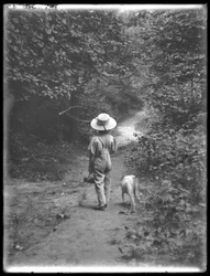 William Gray Hassler w słomkowym kapeluszu i kombinezonie, niosący wędkę i kosz, idąc ścieżką w lesie z małym psem (Bessie), 20 sierpnia 1911