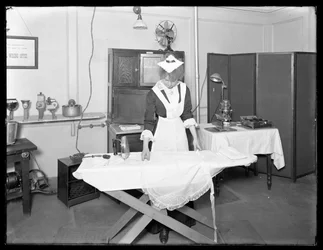 Młoda kobieta w stroju pokojówki demonstruje użycie elektrycznego żelazka, ok. 1913-14